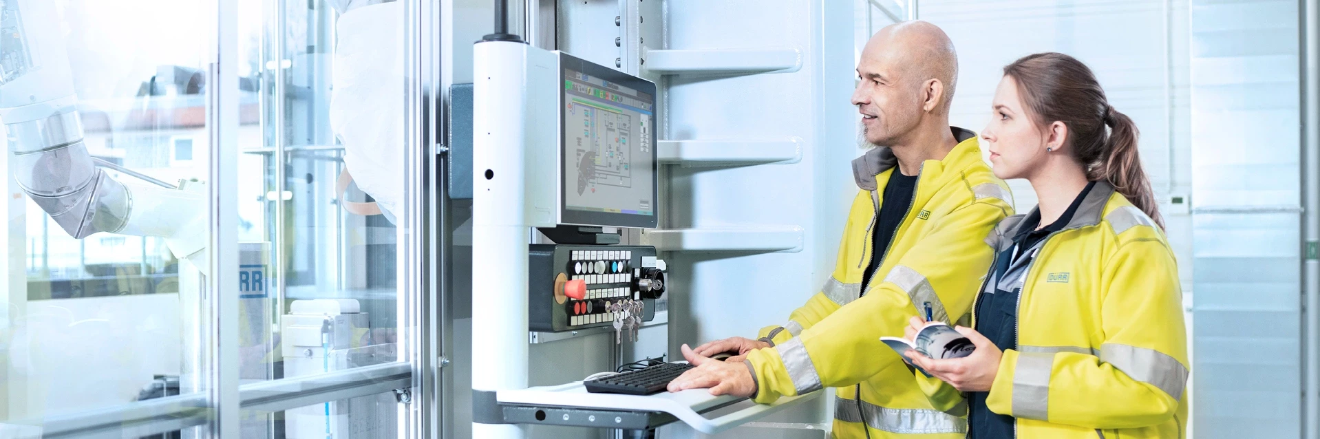 Two technicians operating an control panel, with a robotic arm visible behind glass.